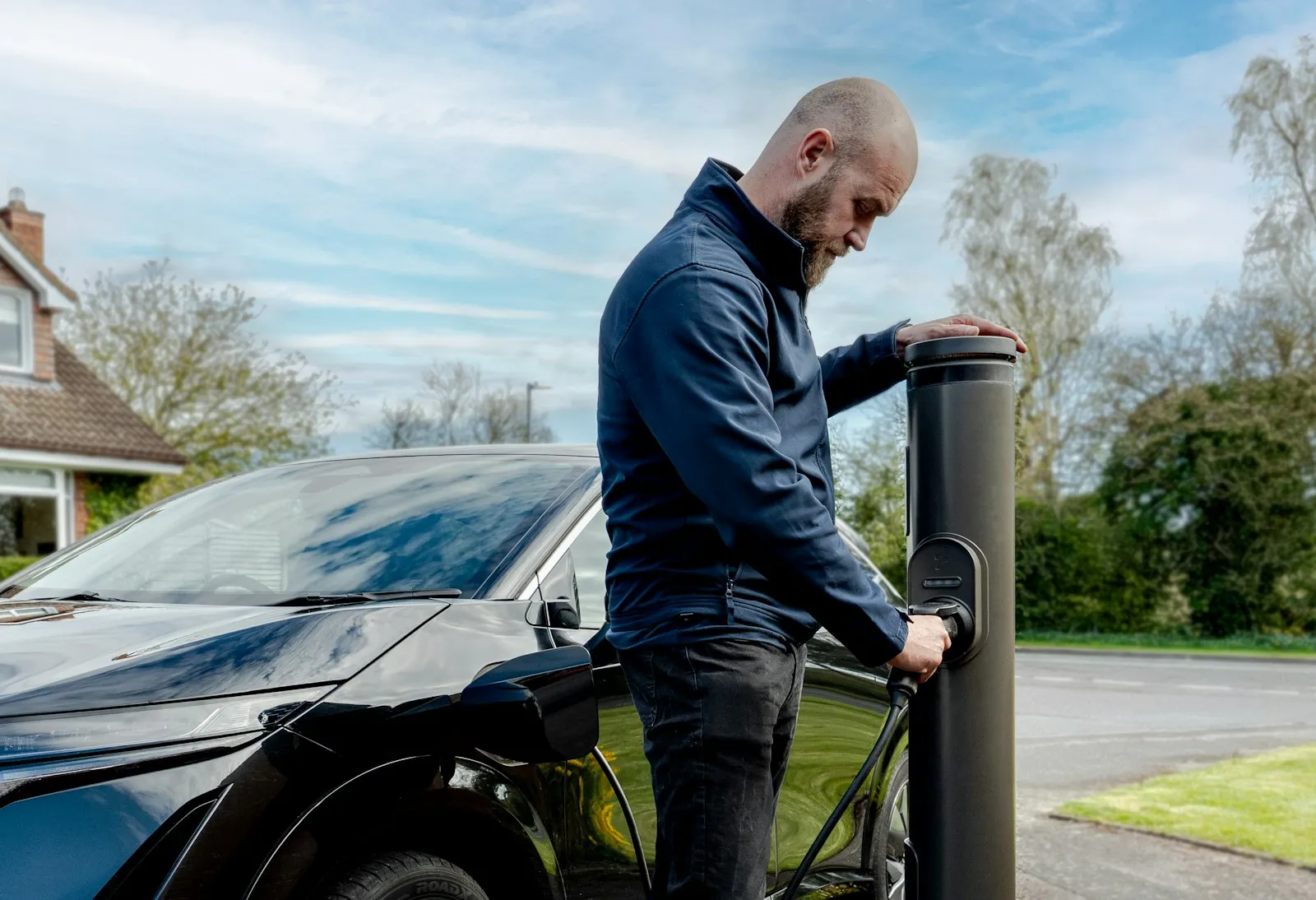 A driver plugging a charging cable into a black electric car at an outdoor charger
