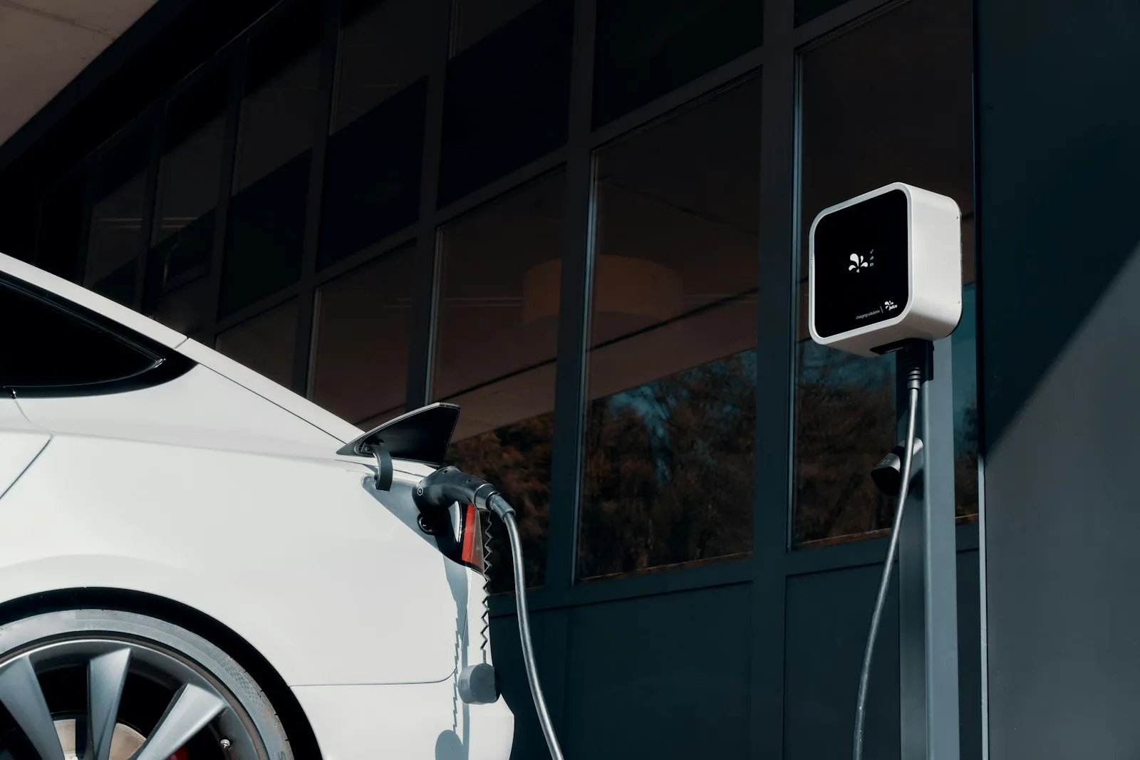 A white electric car plugged into a wall charger outside a modern office building